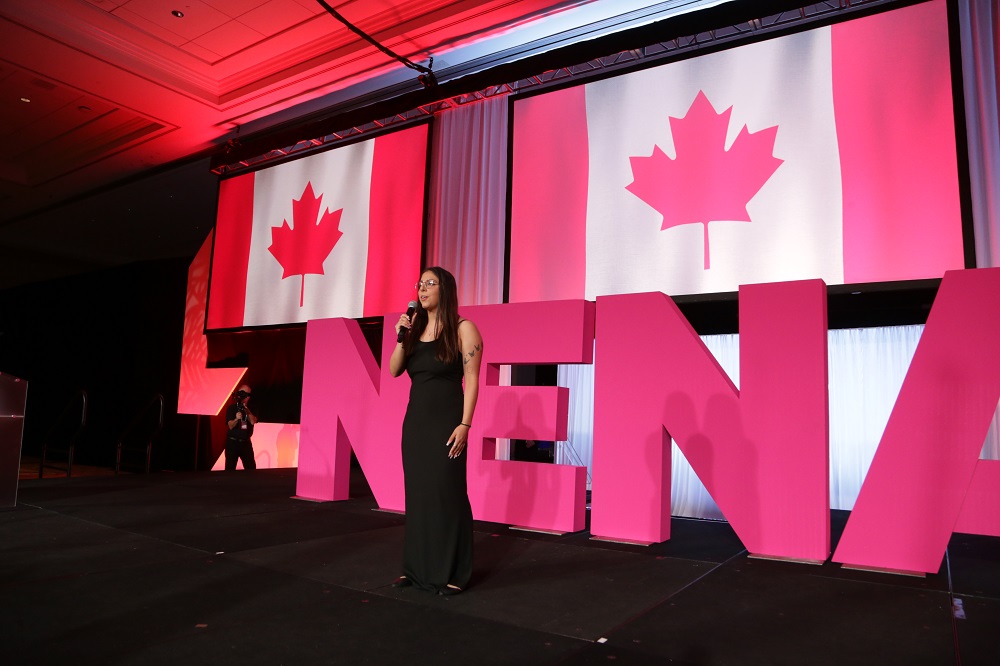 A woman stands on a stage holding a microphone in front of a large sign with the letters N-E-N-A and projection screens with images of the Canadian flag. 