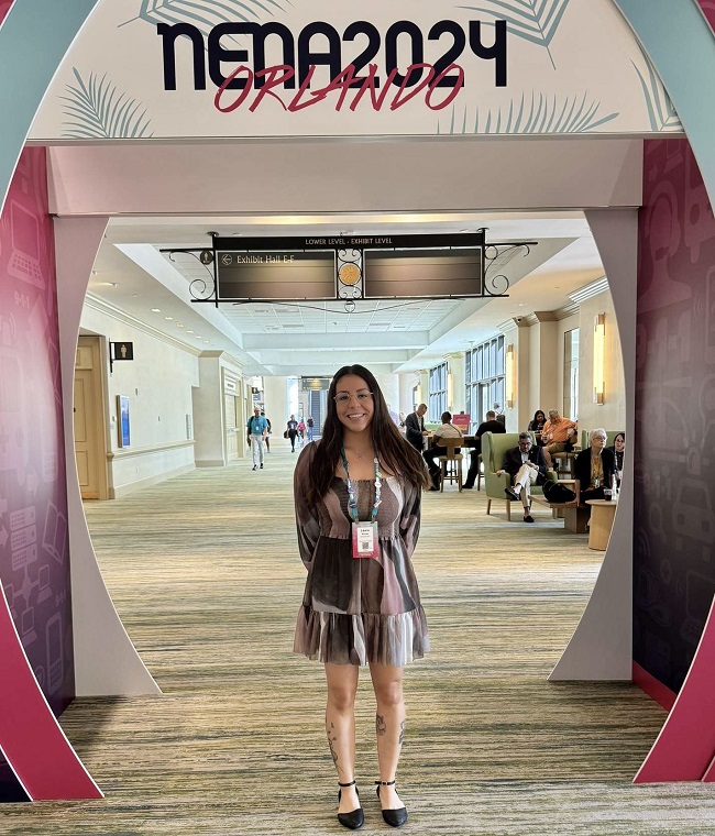 A woman stands smiling below a sign reading "NENA 2024 Orlando". 